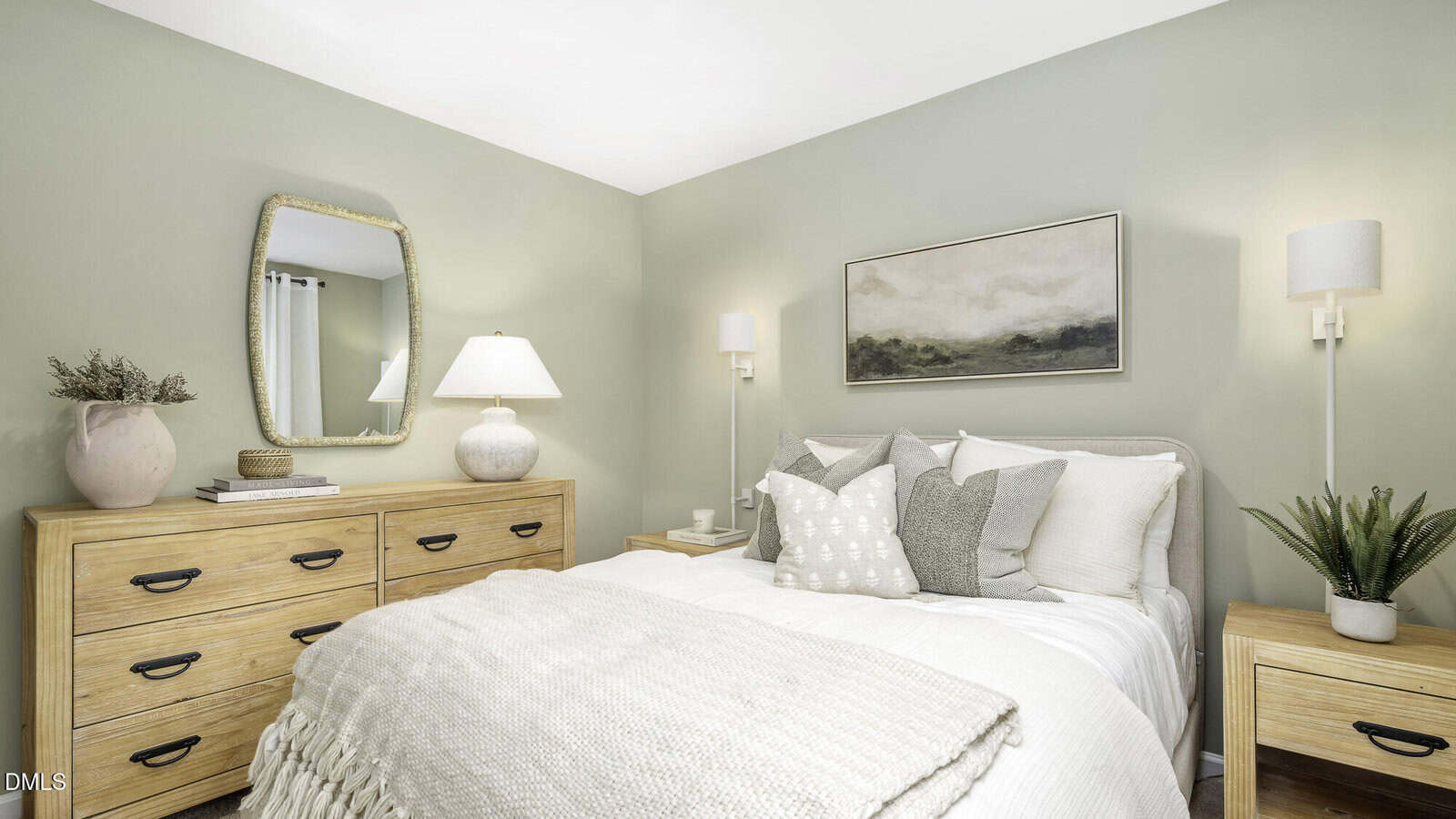 A cozy bedroom with light green walls, a bed with white and beige bedding, two wooden nightstands, a wooden dresser with a lamp and mirror, and wall-mounted lights. A potted plant and framed landscape art decorate the room.