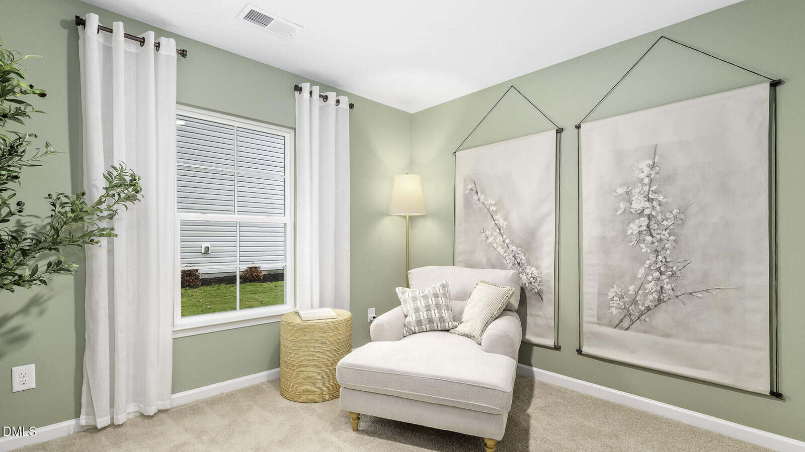 A cozy reading nook with a beige armchair, pillows, a wicker ottoman, and a floor lamp. Light green walls feature two large botanical artworks; a window with white curtains lets in natural light.