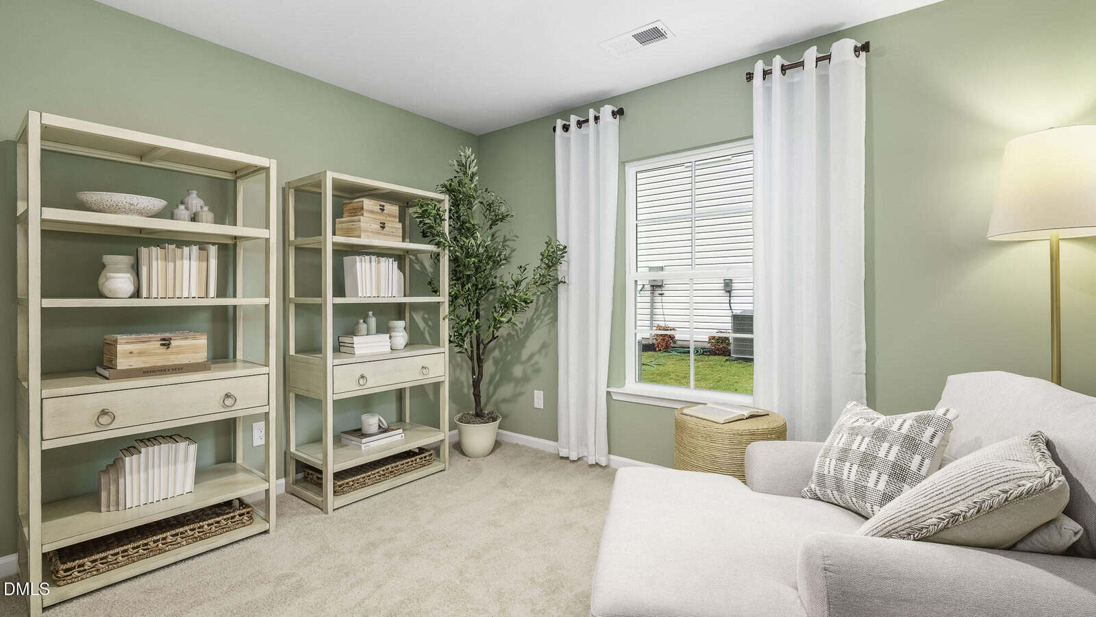 A cozy home office with light green walls, beige carpet, a white armchair with patterned pillows, two wooden bookshelves with books and decor, a potted plant, a wicker basket, and sheer white curtains on a window.