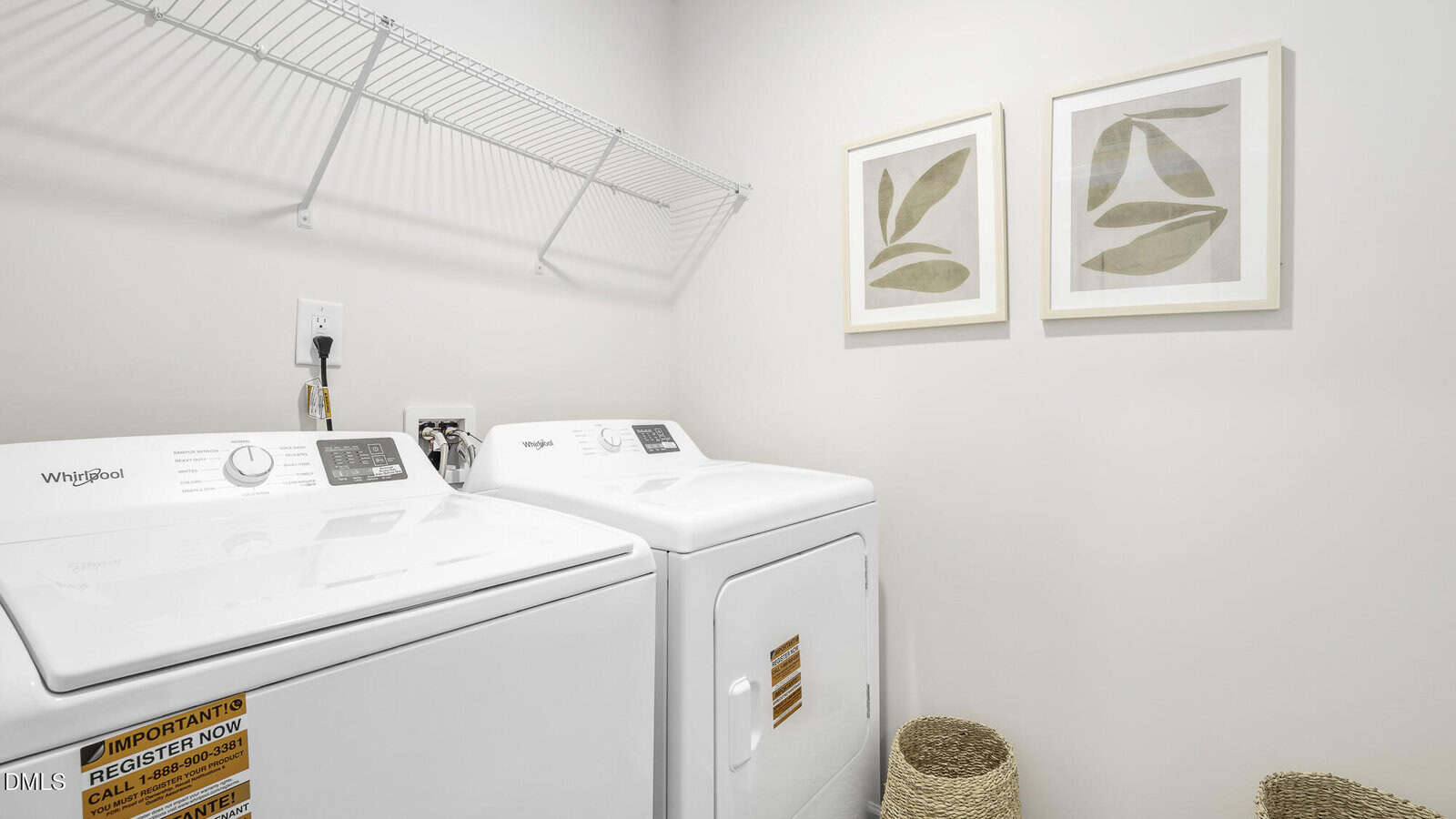 A bright laundry room with a white Whirlpool washer and dryer, two woven baskets on the floor, a wire shelf above the appliances, and two framed abstract leaf prints on the wall.