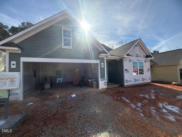 A newly constructed house with green siding, an unfinished garage, and a "SOLD" sign in the window. Construction materials are visible inside the garage, and sunlight shines from above. The ground is covered in red dirt and gravel.