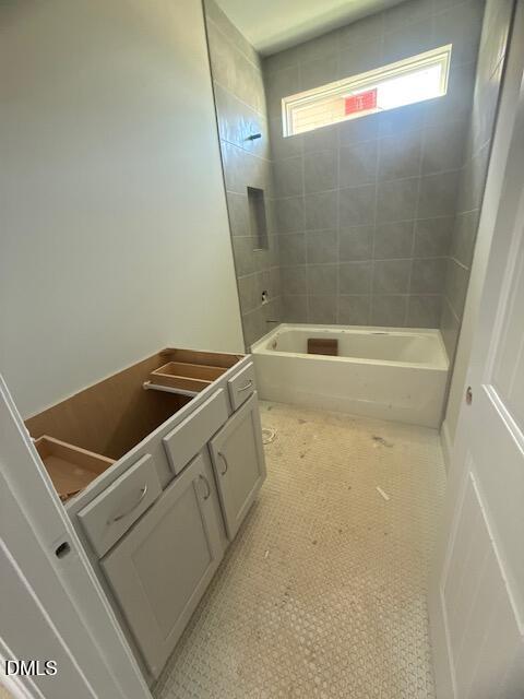 A bathroom under construction with an unfinished vanity, no sink or countertop, a white bathtub with gray tiled walls, hexagonal floor tiles, and a small high window letting in natural light.