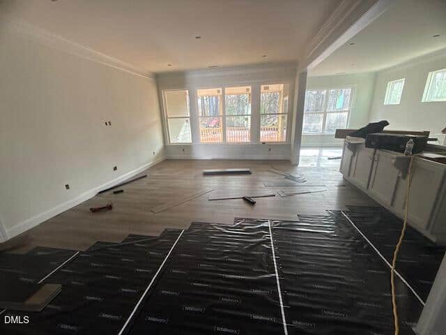 A bright, unfurnished room with large windows and partially installed wood flooring. Installation tools and materials are scattered on the floor, and the kitchen area is visible to the right.