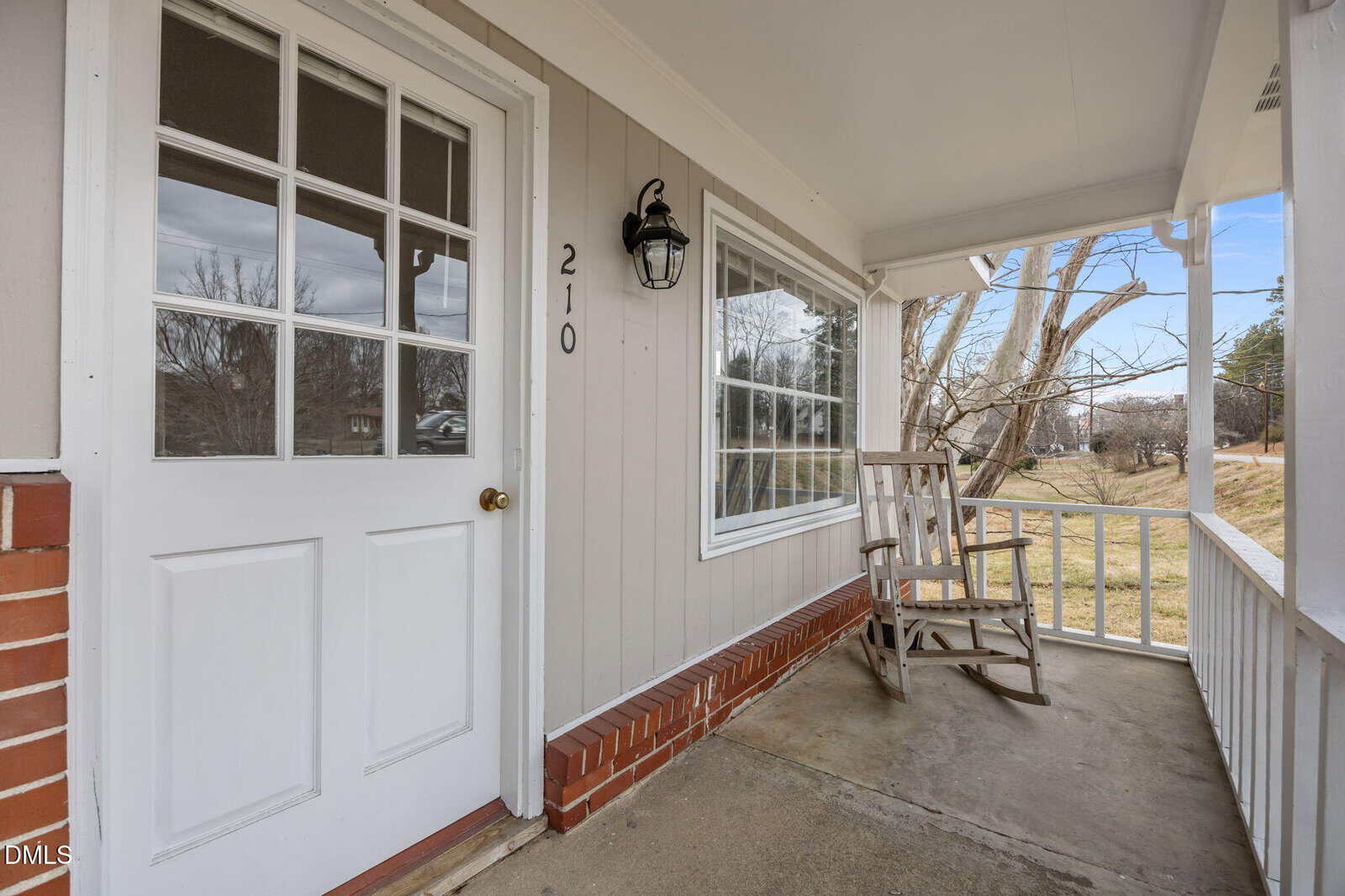 A covered front porch with a white door numbered 210, a large window, and a wooden rocking chair. The porch has a white railing and overlooks a grassy yard with trees.