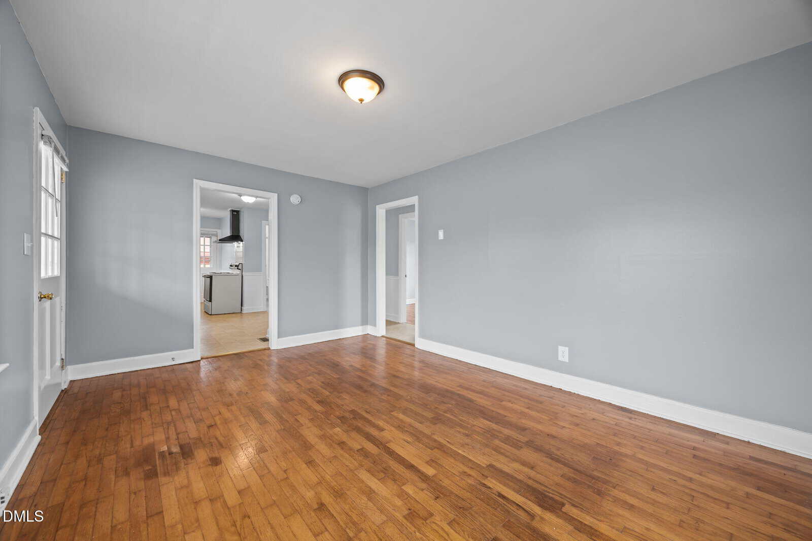 Empty room with light gray walls, wooden floor, white trim, single ceiling light, and doorways leading to other rooms. The kitchen is partially visible through an open doorway in the background.