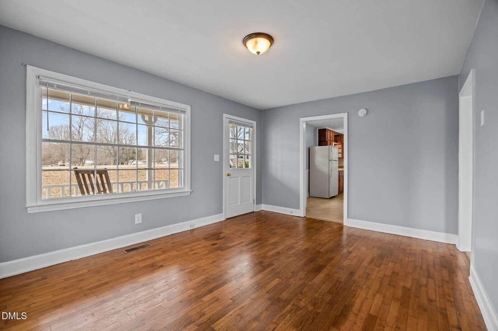 A bright room with hardwood floors, gray walls, a ceiling light, a large window, a white door, and an open doorway leading to a kitchen with wooden cabinets. Outdoor scenery is visible through the window.