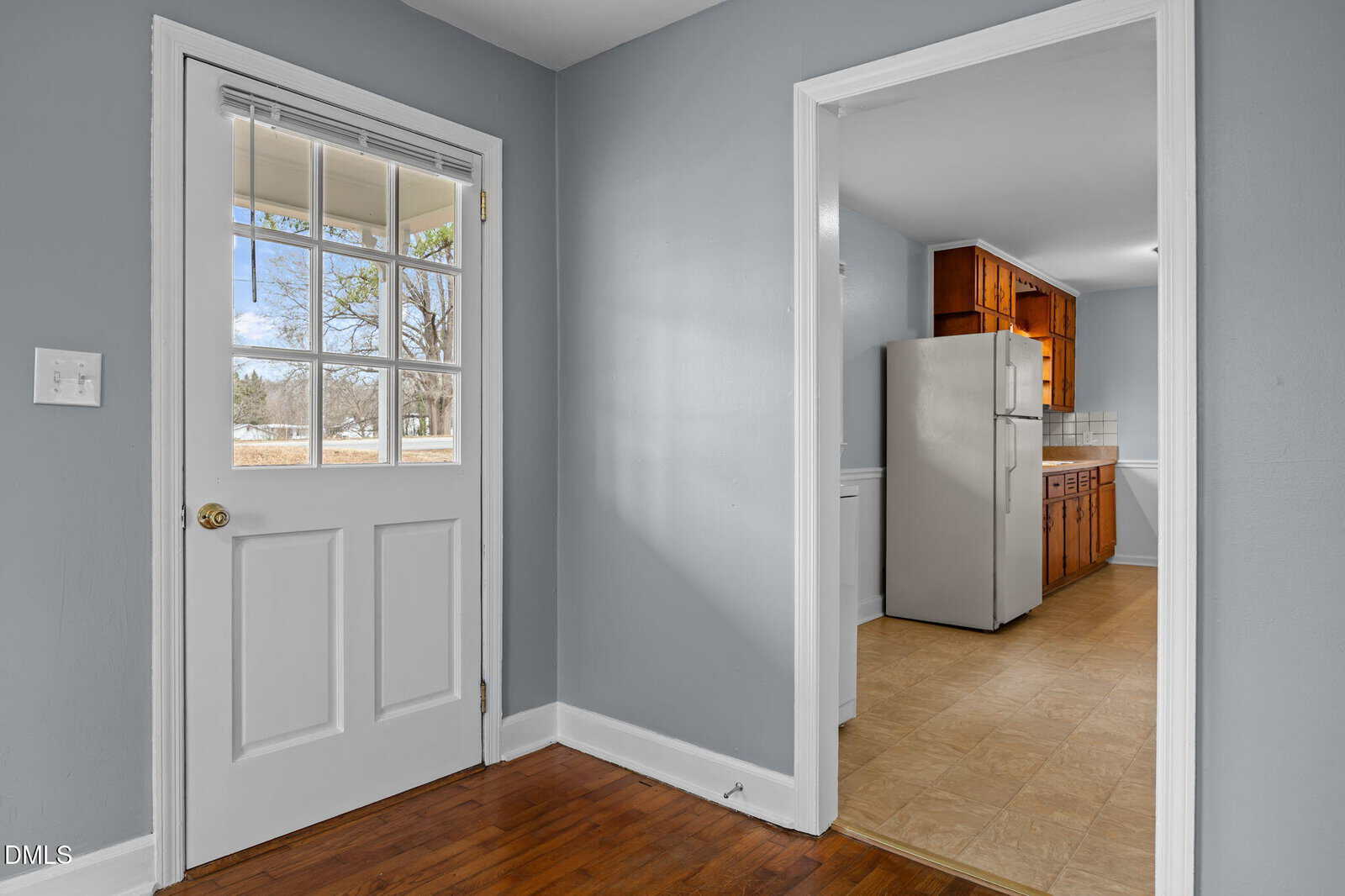 A small entry area with a glass-paned door, wood floor, and light gray walls opens to a kitchen with wood cabinets, a refrigerator, and beige tile flooring. Trees are visible through the door window.