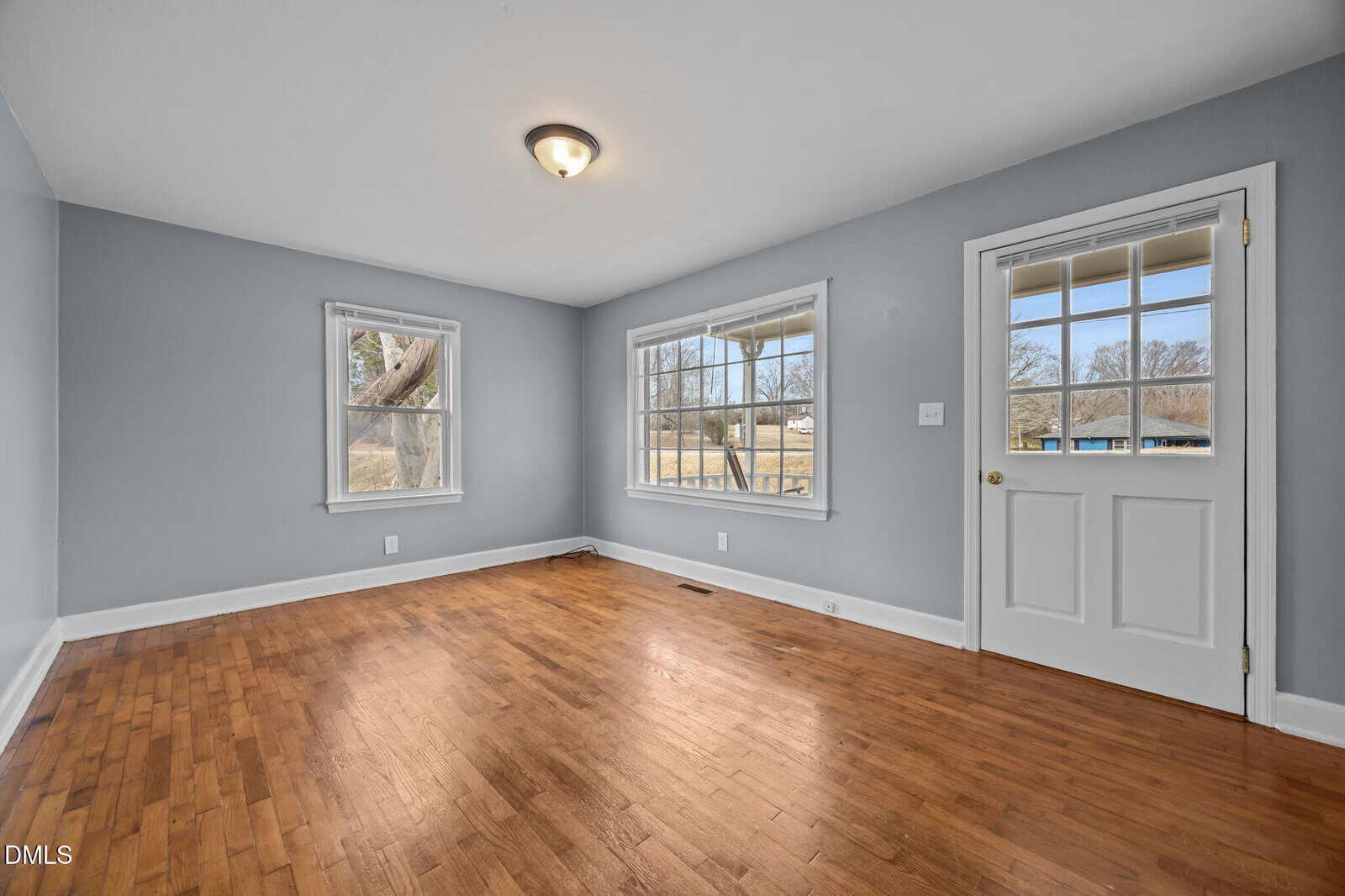 A bright, empty room with light blue-gray walls, hardwood floors, two large windows, and a glass-paneled door. Natural light fills the space, and tree branches are visible outside.