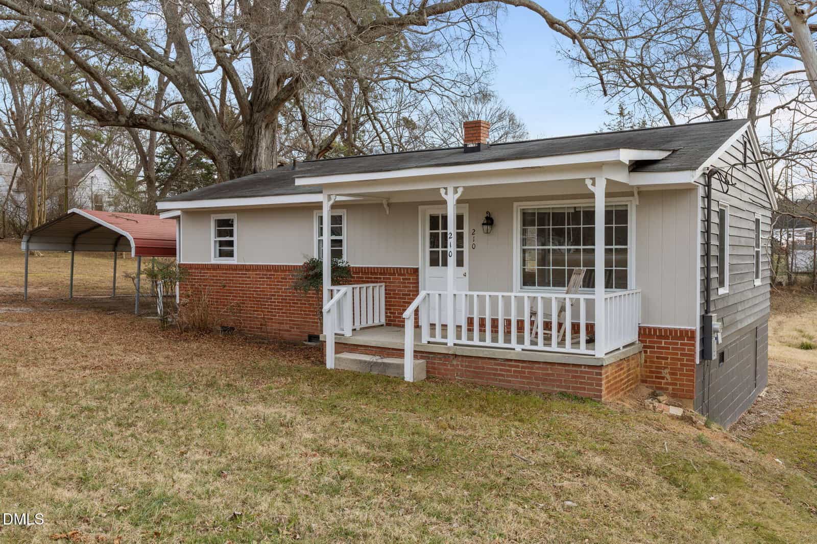 A small, single-story house with gray siding, red brick accents, a white front porch, and a sloped roof. There's a carport to the left and large trees in the yard on a slightly sloped grassy lot.