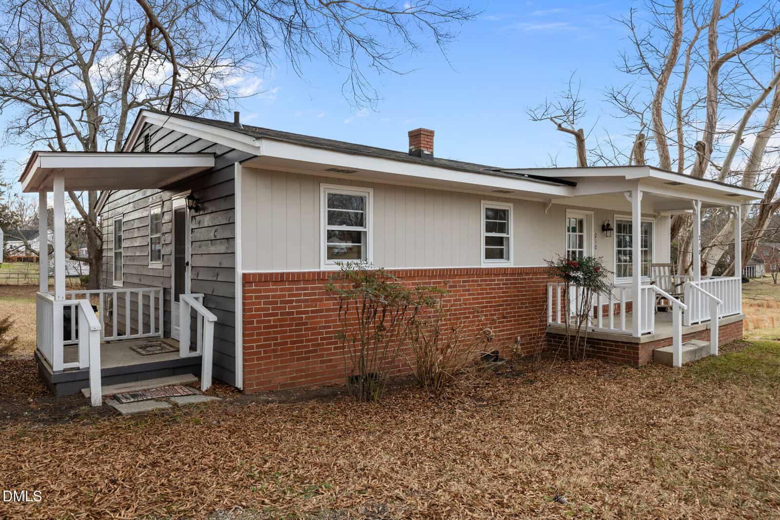 Single-story house with brick and wood siding, covered front and side porches, white railings, and leaf-covered yard; leafless trees and blue sky in the background.