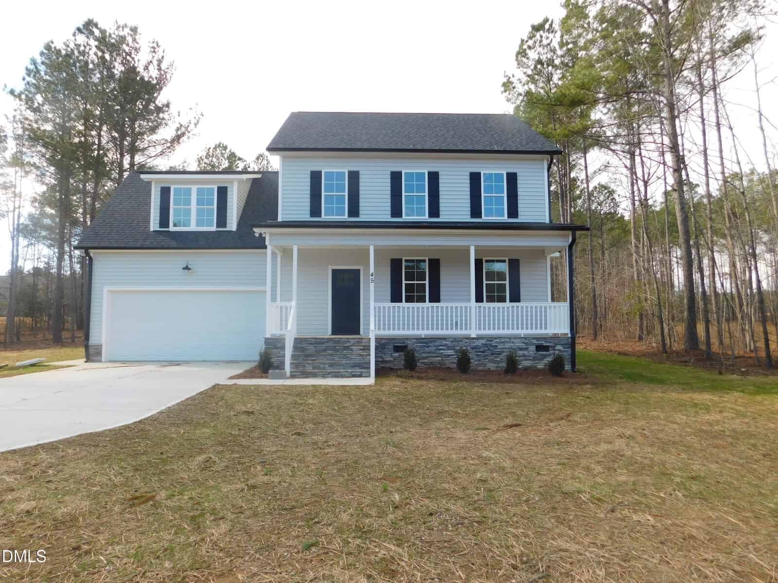 Two-story house with light gray siding, black shutters, and a covered front porch at 45 Copper Penny Road. The attached two-car garage is on the left. This charming Zebulon home sits on a grassy lot bordered by tall trees, with a concrete driveway leading to the garage.