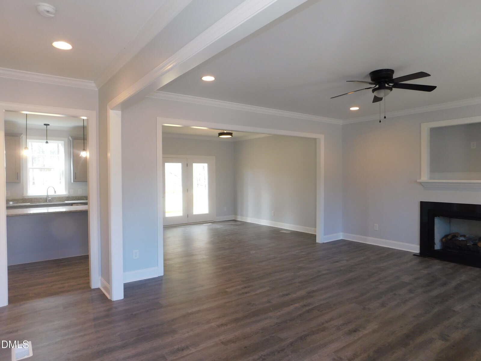 Spacious open-concept living area with gray wood flooring, light gray walls, a black ceiling fan, recessed lighting, a fireplace, and large windows. Kitchen with stainless steel sink is visible through a wide doorway.