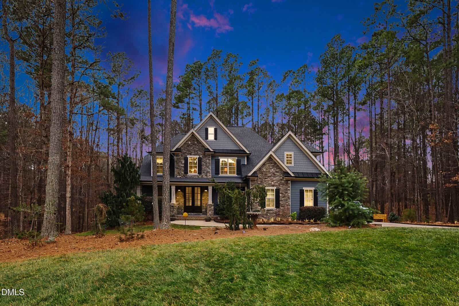 Large, two-story house with stone and siding exterior, lit windows, and a front porch, surrounded by tall trees and a grassy yard at dusk with a colorful sky.