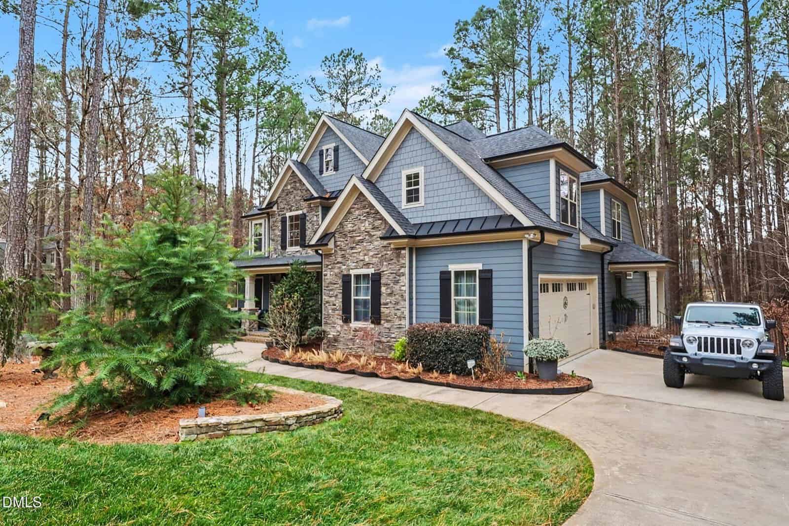 A two-story blue house with stone accents, white trim, and a gray roof sits beside a wooded area. A white jeep is parked in the driveway, and the yard is landscaped with shrubs, mulch, and a small pine tree.