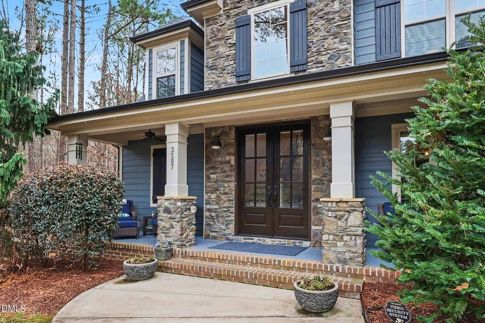 Two-story house with stone accents, blue siding, black shutters, and a covered front porch with columns. There are large double doors, shrubbery, and two potted plants by the entrance.