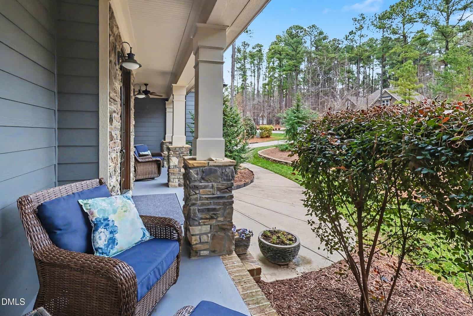 Wicker chairs with blue cushions sit on a stone porch overlooking a landscaped yard, walkway, and trees under a blue sky.