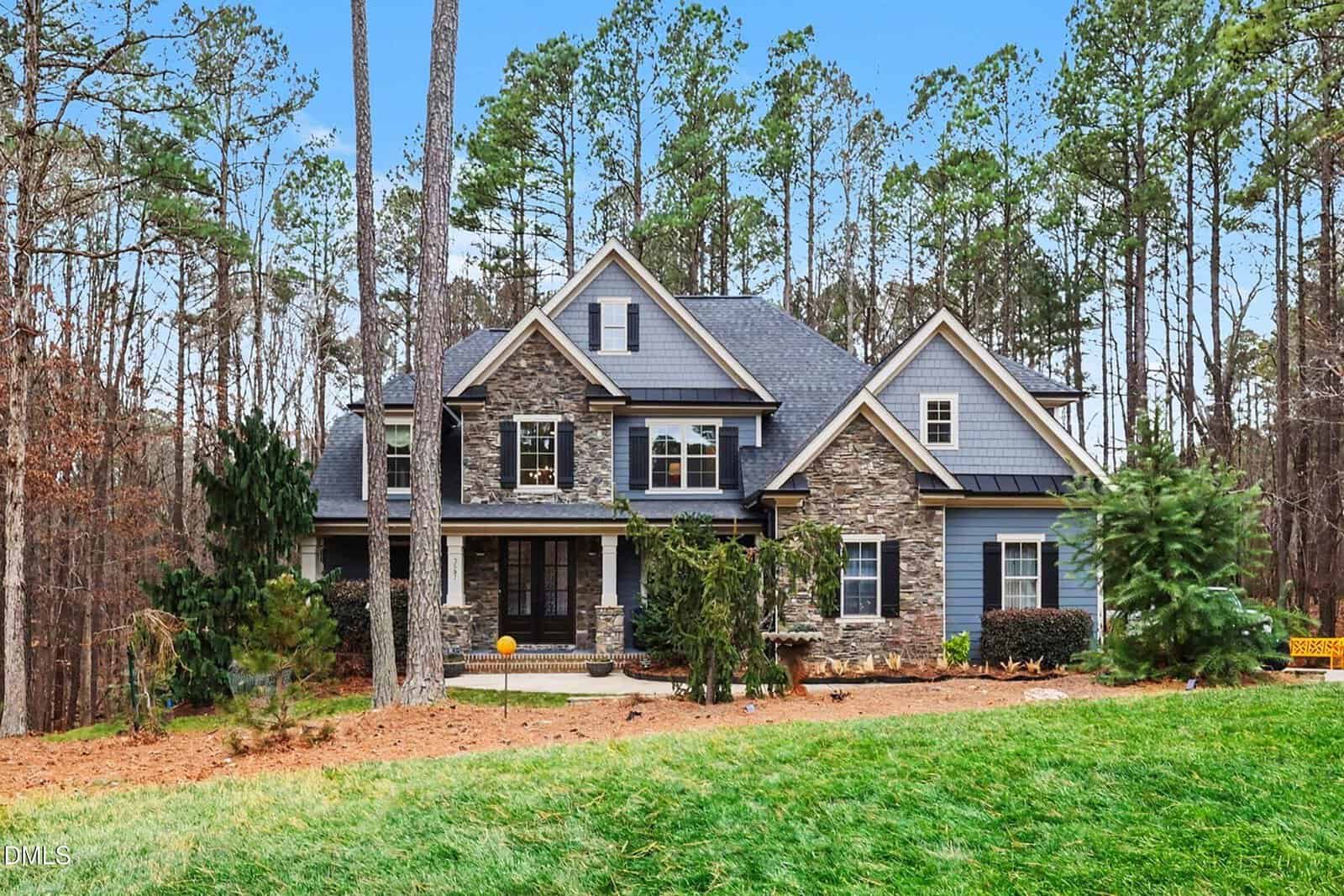 Charming two-story house at 3587 Garner Terrace Way, Wake Forest, with stone and blue siding, gabled roof, and multiple windows, nestled among tall pine trees and landscaped lawn with shrubs and mulch under a clear blue sky.