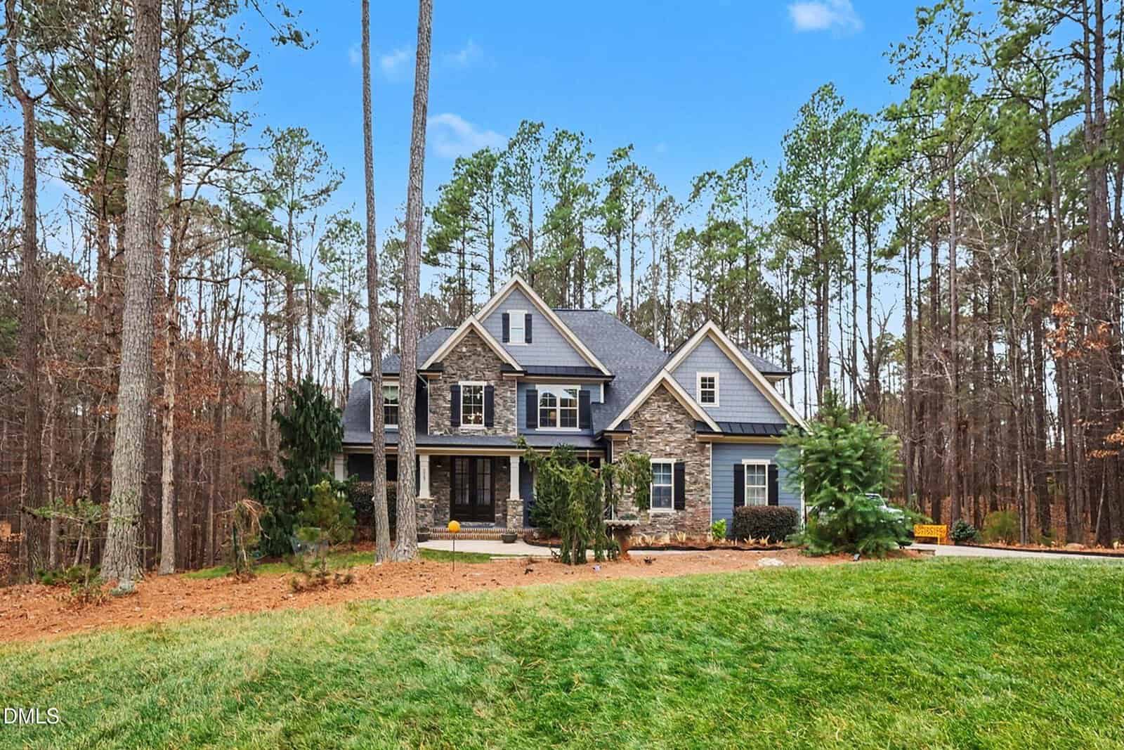 Two-story house with stone and wood exterior set among tall pine trees, featuring a large front lawn, a covered porch, and a paved driveway on a clear day.