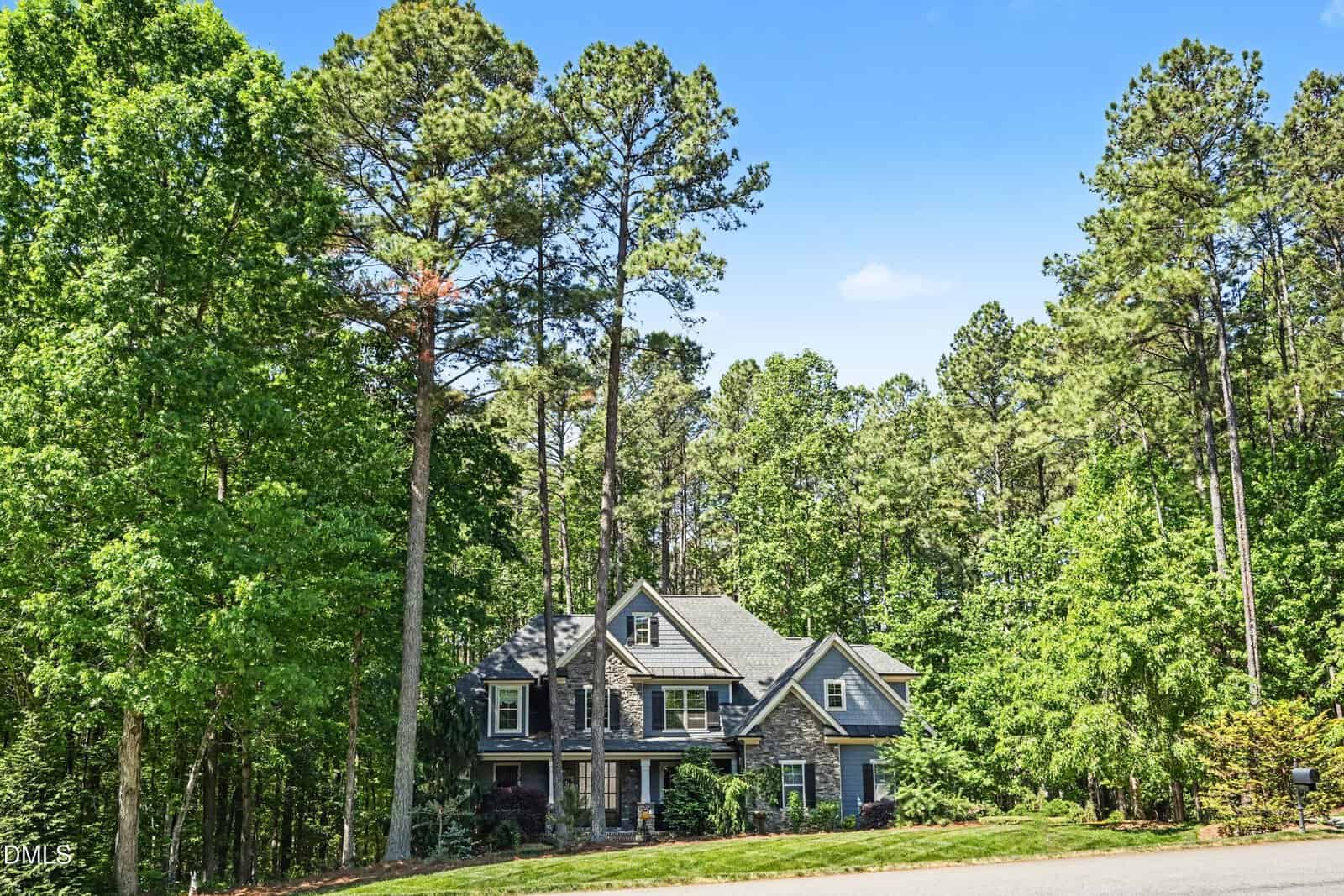 A two-story gray house with stone accents sits among tall pine trees and lush green foliage under a clear blue sky, viewed from a quiet street.
