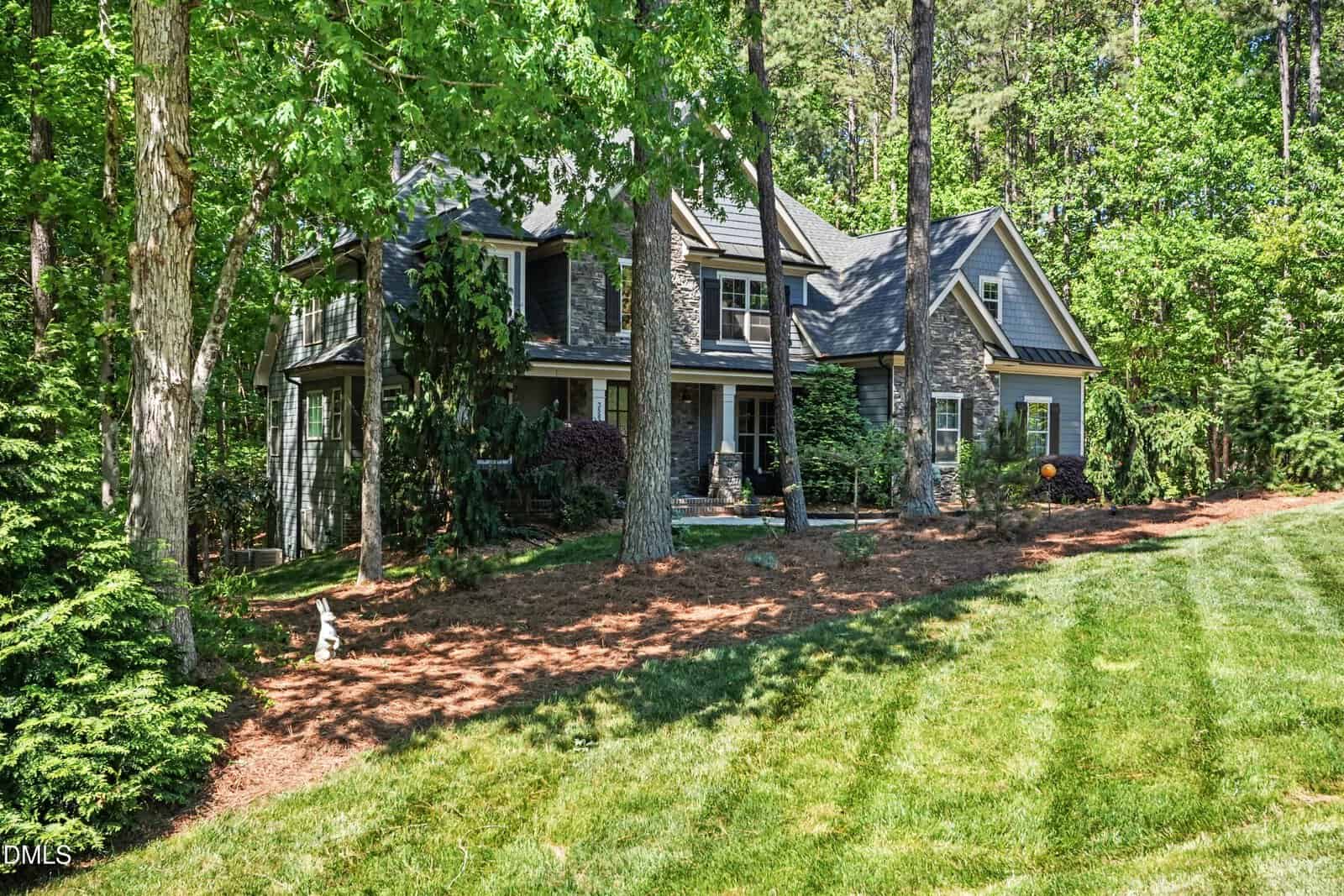 A large two-story house with gray siding and stone accents sits surrounded by tall trees and lush greenery, with a well-maintained lawn sloping in front of the home.