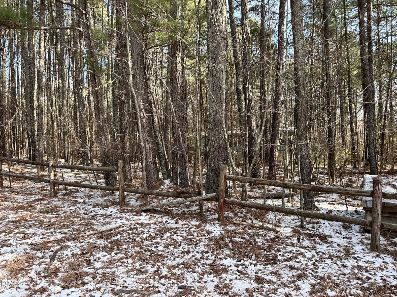 A wooden fence with broken sections runs through a snowy, wooded area near 2160 Frazier Lane in Franklinton, where tall trees and patches of brown leaves cover the ground as sunlight filters through bare branches.