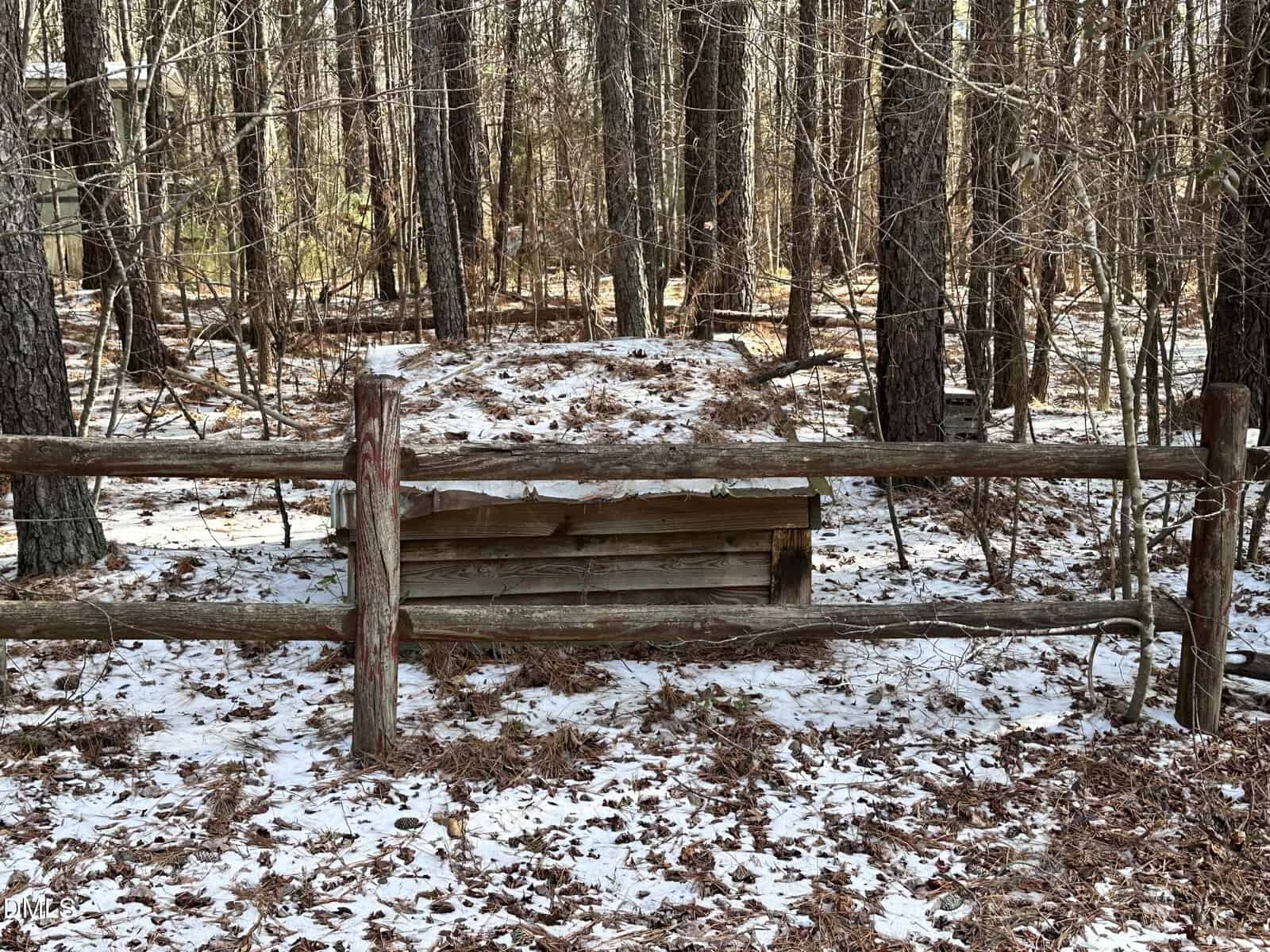 A wooden structure covered with snow sits behind a rustic wooden fence in a forest with tall trees and a light dusting of snow on the ground.