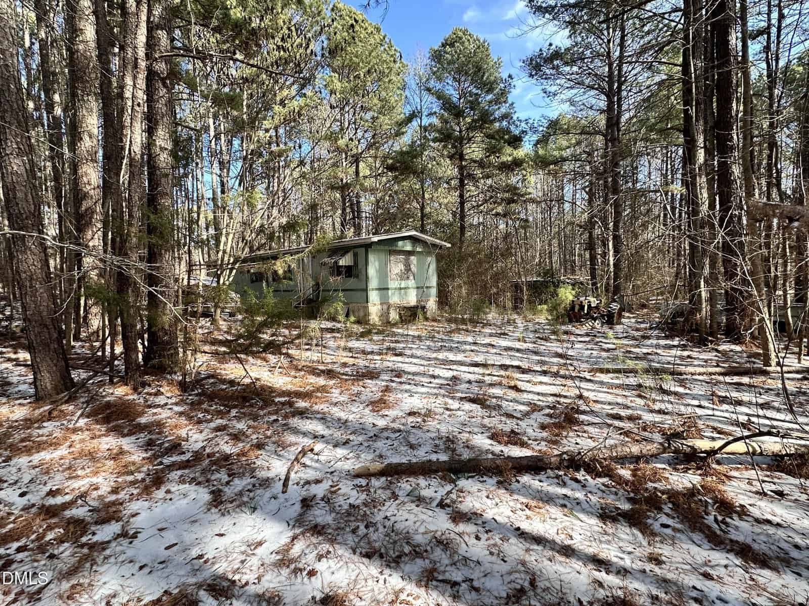 A green, weathered mobile home sits among tall pine trees in a forest. The ground is lightly covered with patches of snow and scattered pine needles under a partly cloudy sky.