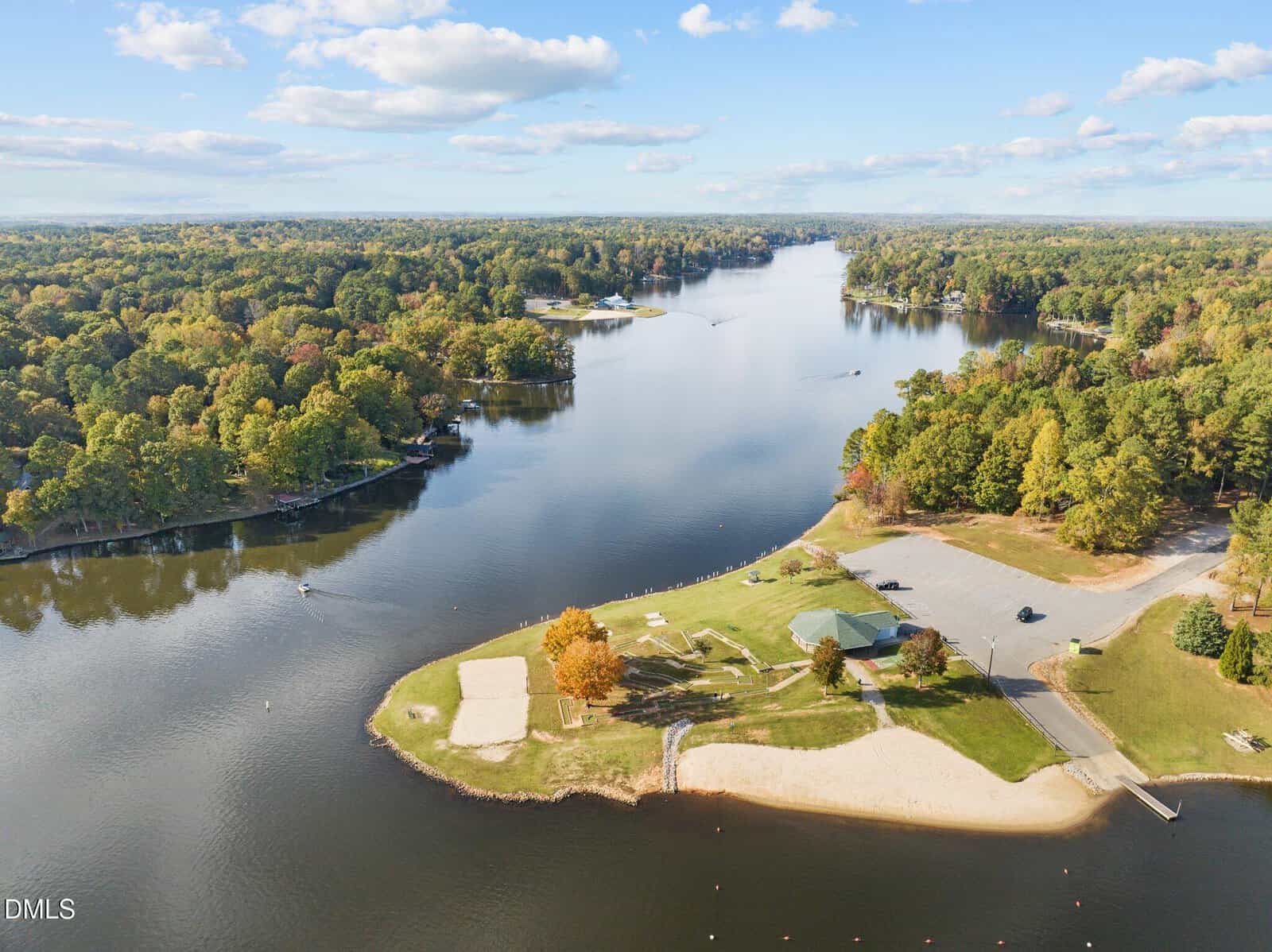 Aerial view of a riverside park with sandy beaches, green grass, a pavilion, parked cars, and forested areas surrounding the river under a partly cloudy sky.