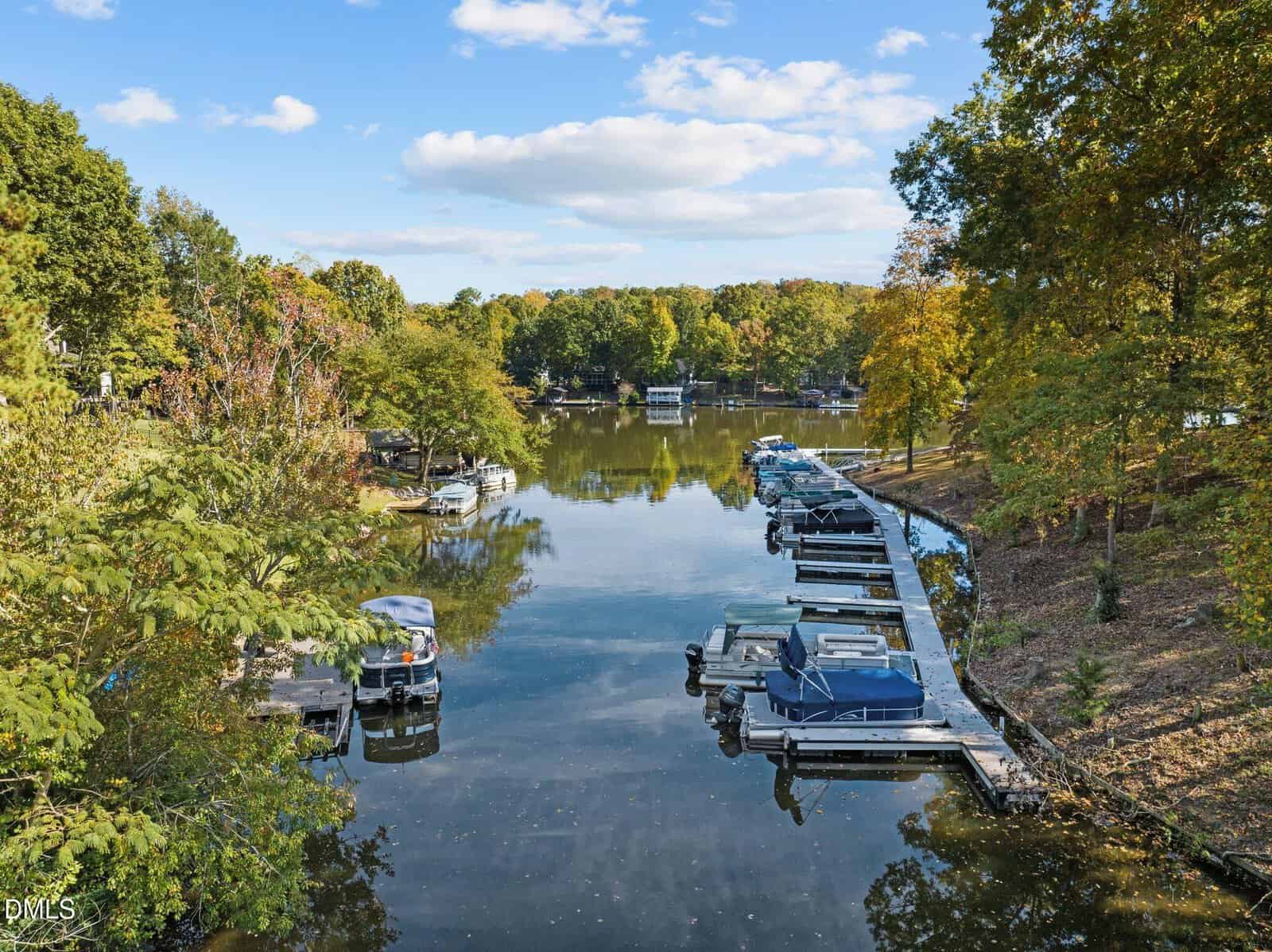 A calm lake with several boats docked along both shores, surrounded by trees with green and yellow foliage under a partly cloudy sky. The water reflects the scenery.