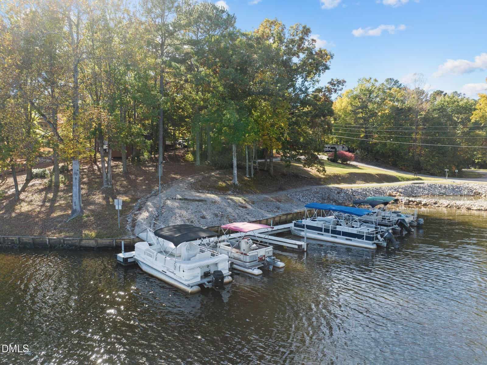 Several pontoon boats are docked at a small marina along a wooded shoreline with a paved road and cars visible in the background under a partly cloudy sky.