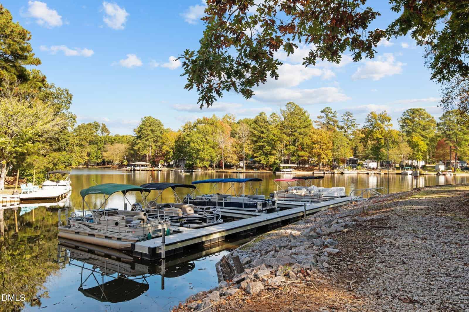 A small marina with several covered pontoon boats docked beside a rocky shoreline, surrounded by green trees and calm water under a blue sky with scattered clouds.