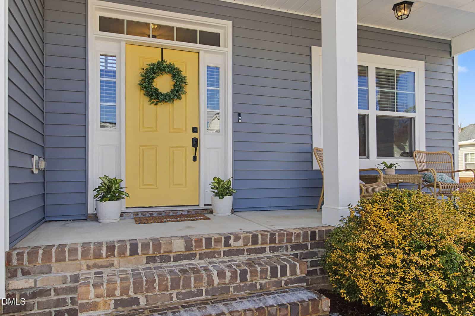 A yellow front door with a wreath is flanked by potted plants on a brick porch. To the right, two wicker chairs and a table sit near a window. Blue-gray siding and a shrub complete the inviting entryway.