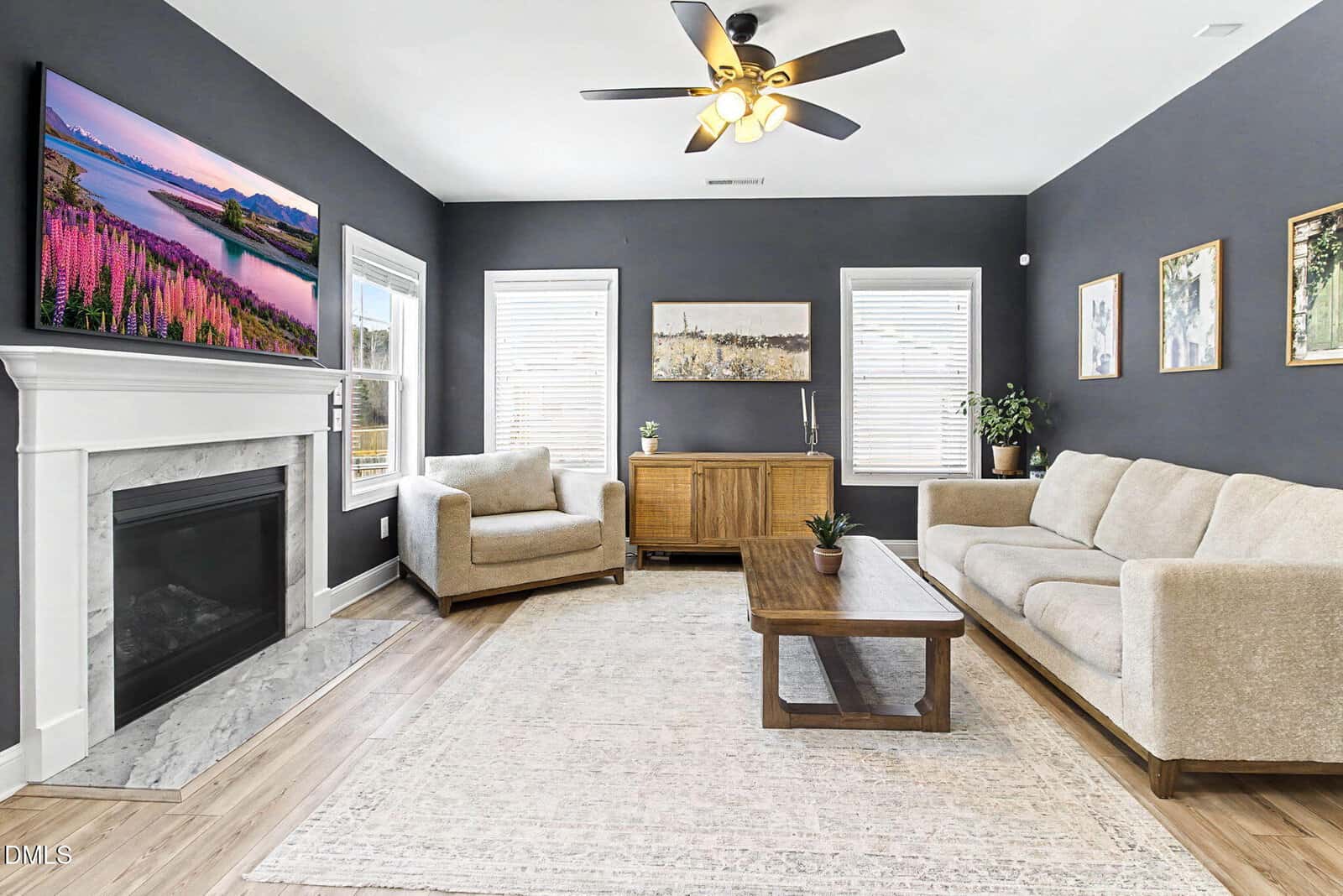 A modern living room with dark gray walls, beige sofas, a wooden coffee table, a white fireplace with a TV above it, large windows with blinds, artwork on the walls, and a ceiling fan with lights.