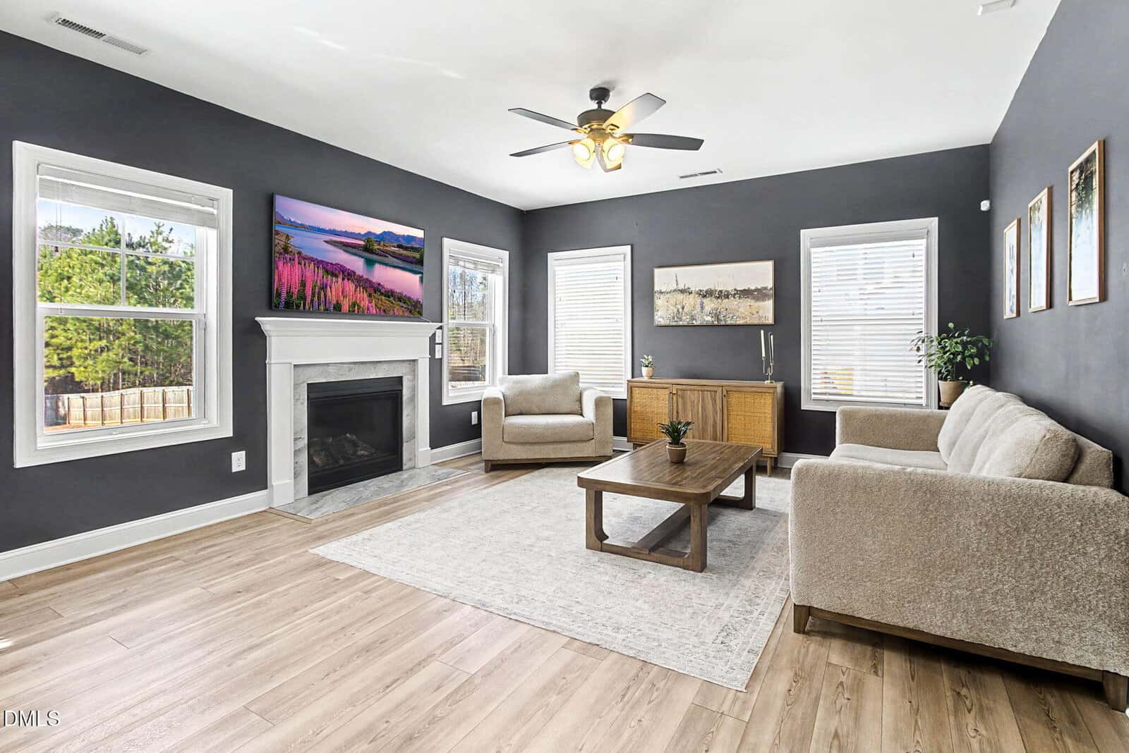 Modern living room with dark walls, light wood flooring, a white fireplace, beige sofas and chairs, a wooden coffee table, and large windows letting in natural light. Art and plants add decor. Ceiling fan overhead.