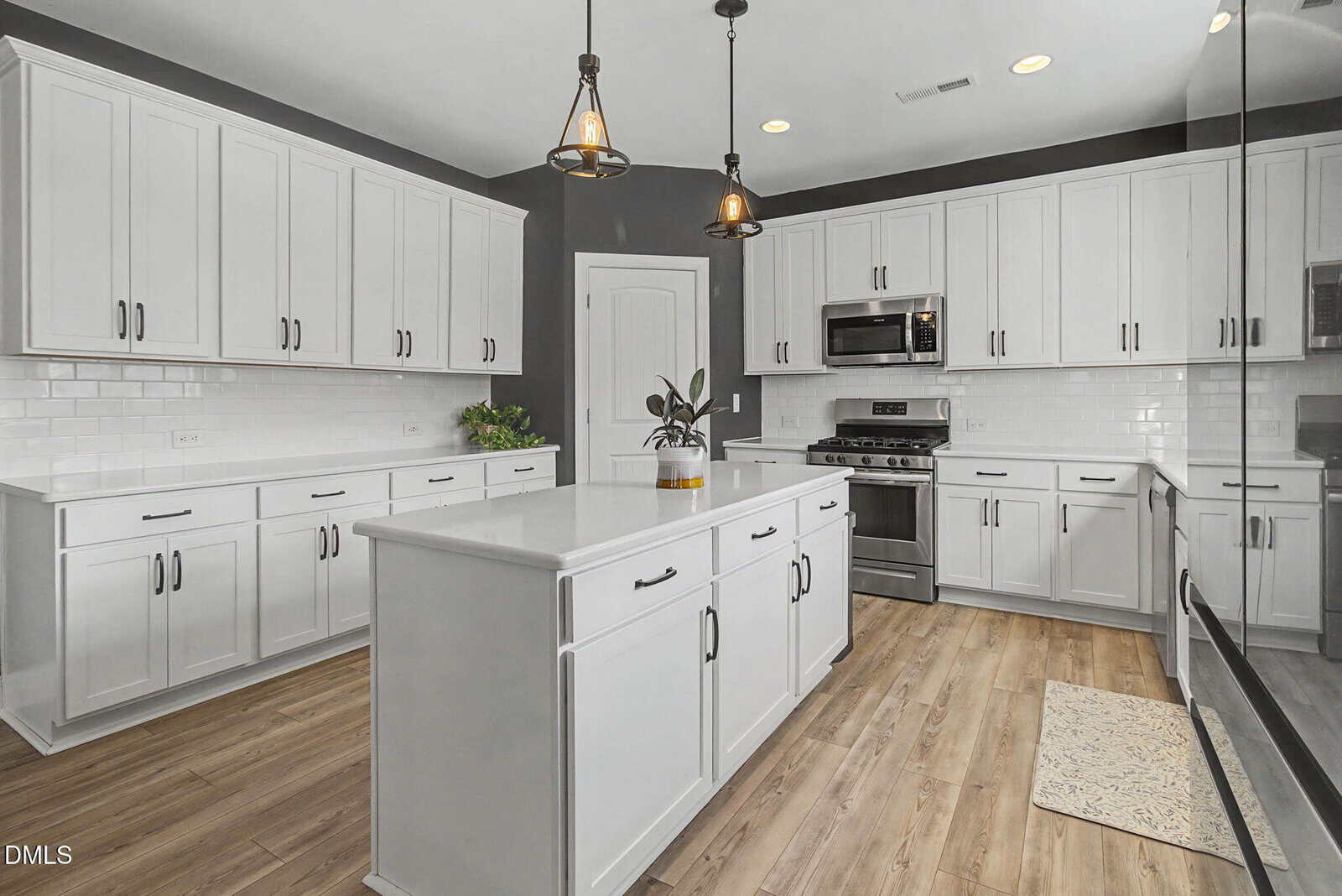 Modern kitchen with white cabinets, stainless steel appliances, a central island, pendant lights, subway tile backsplash, wood flooring, and a small plant on the island. A door and a floor mat are visible.