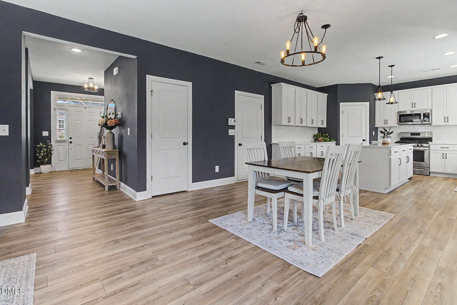 Modern kitchen and dining area with white cabinets, stainless steel appliances, a wooden dining table with four chairs, light wood flooring, and gray walls; entryway visible in the background.