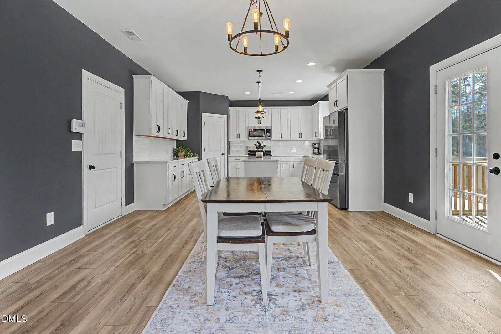 Modern kitchen with gray walls, white cabinets, stainless steel appliances, a wooden dining table with four chairs, light wood flooring, a rug under the table, and a glass door leading outside.