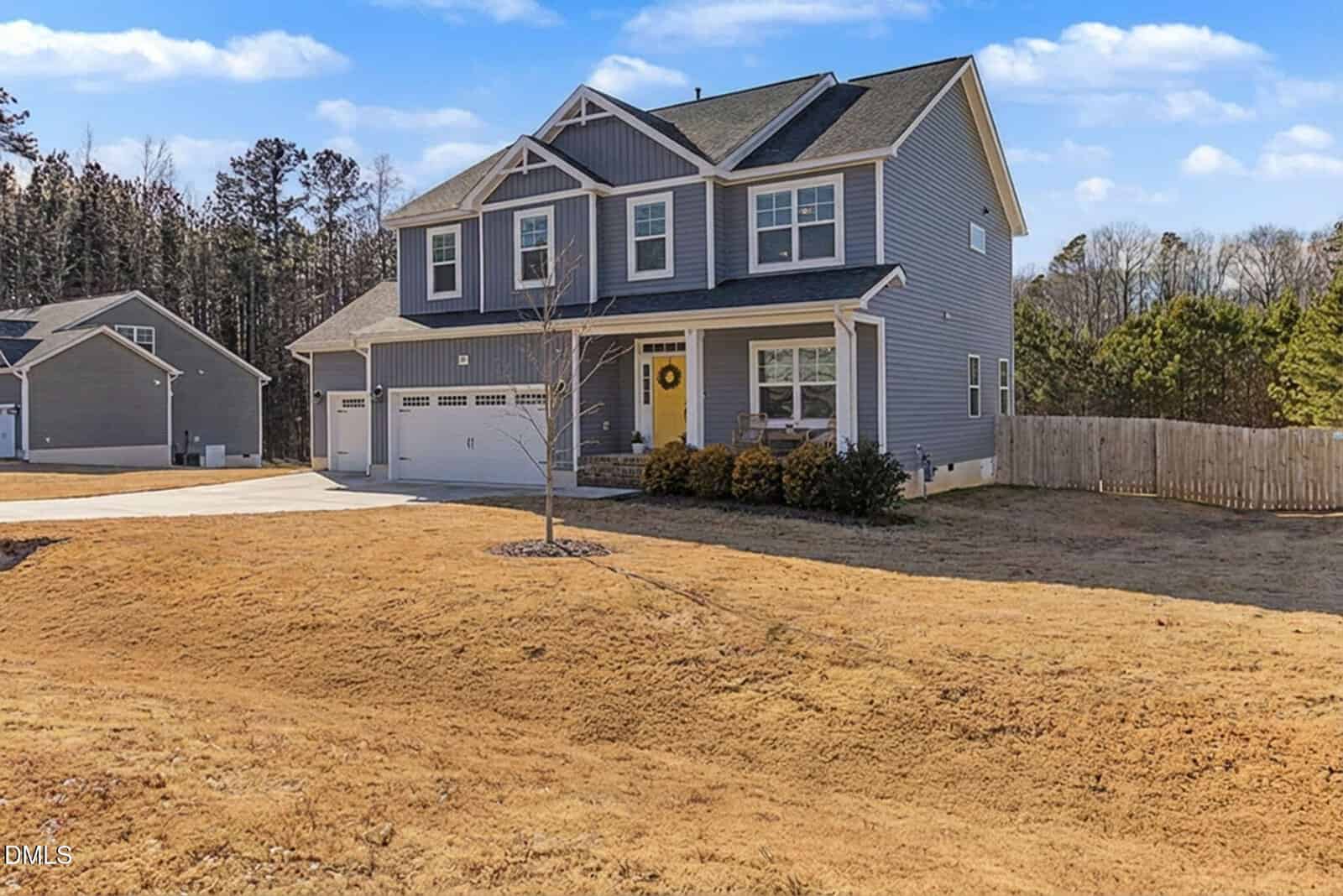 A two-story gray house with a front porch, three-car garage, and a small tree in the yard, surrounded by a wooden fence and dry grass under a blue sky with scattered clouds.
