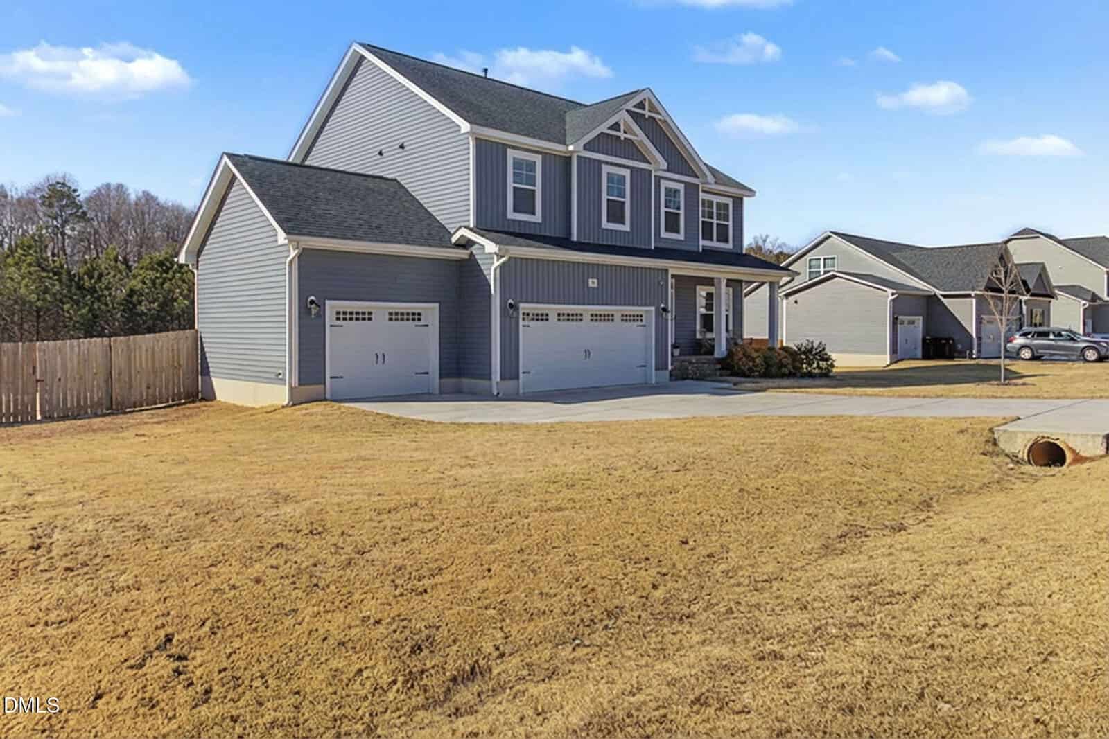 Two-story gray house with white trim, three-car garage, and a large driveway. The house sits on a spacious, grassy yard with a wooden fence on one side and neighboring houses in the background under a blue sky.