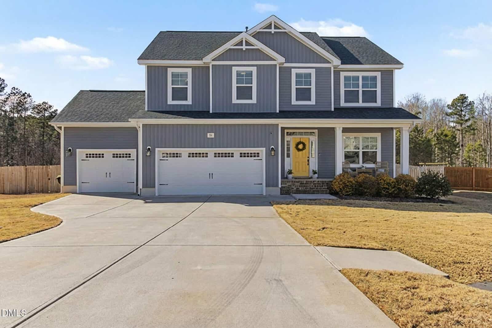 Two-story gray house with white trim, a yellow front door, three-car garage, covered front porch, large driveway, and well-maintained lawn, surrounded by a wooden fence and trees in the background.