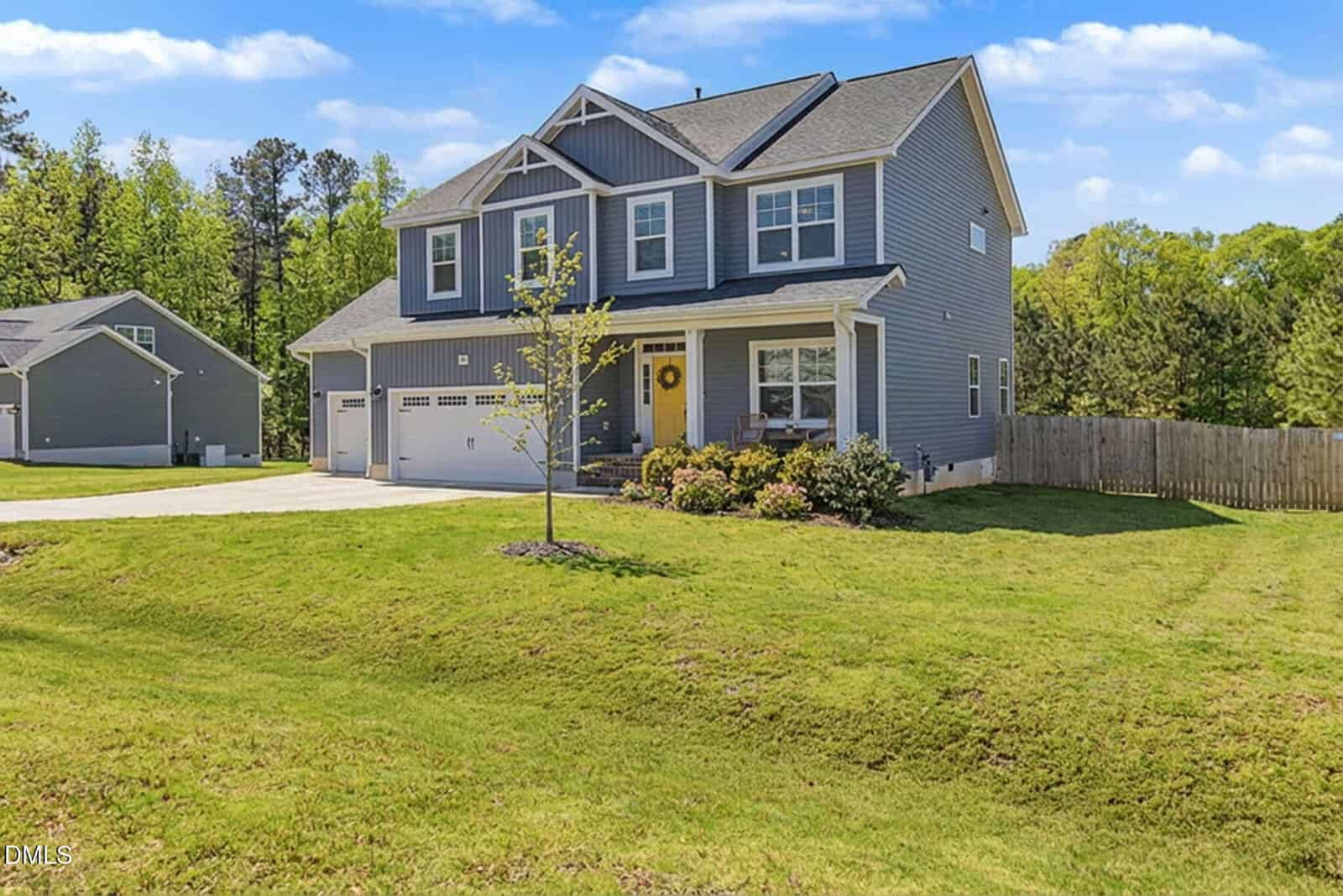 A two-story gray house with white trim, a front porch, and a three-car garage sits on a large, well-manicured lawn, with a young tree and shrubs in the front and a wooded area in the background.