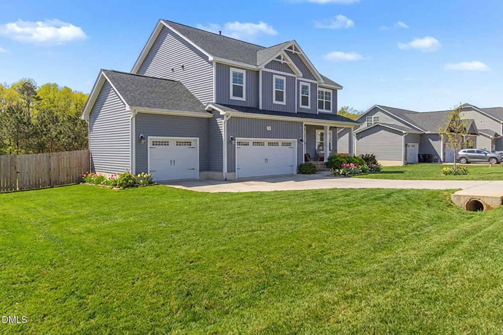 A two-story gray suburban house with white trim, a three-car garage, and a well-maintained green lawn, set on a sunny day with blue skies and neighboring homes visible in the background.