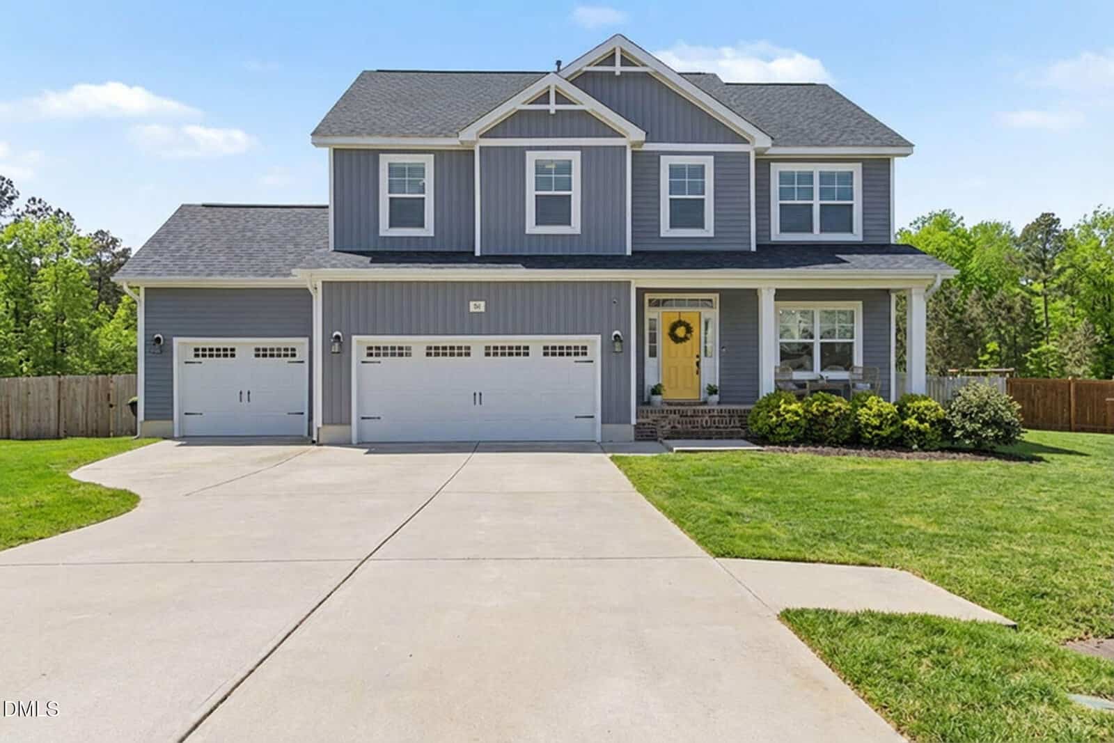 Two-story gray suburban house with white trim, three-car garage, yellow front door with wreath, large driveway, green lawn, and trees in the background under a blue sky.