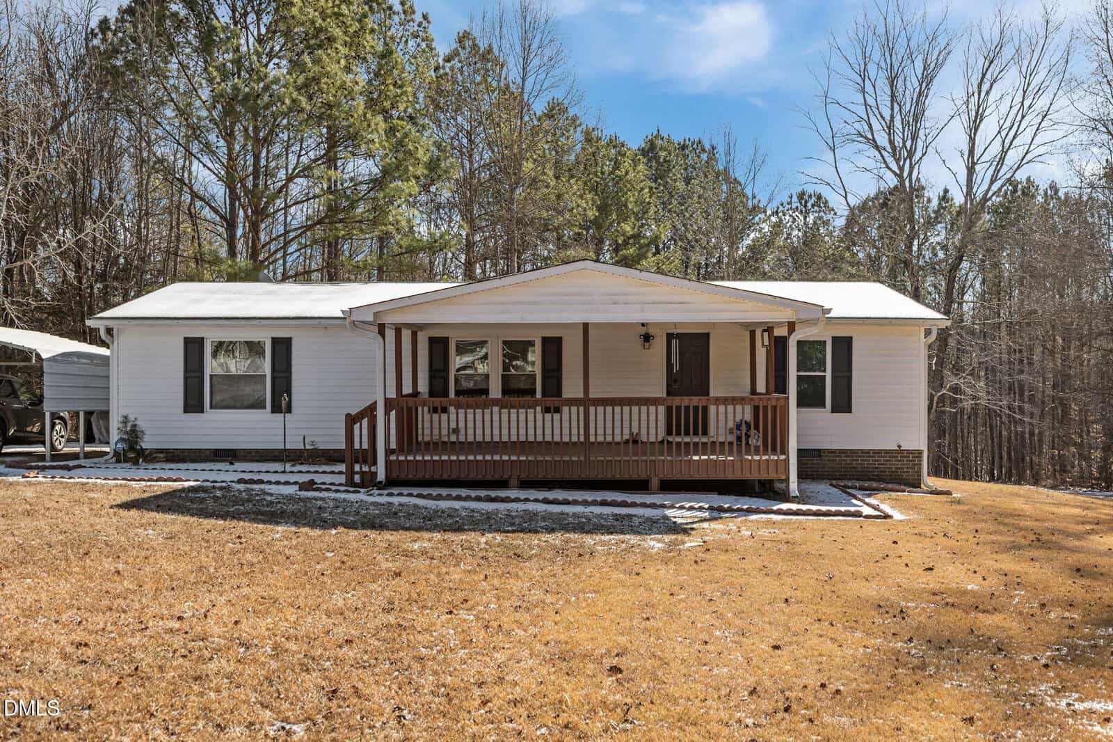 A single-story white house at 1700 Mineral Springs Drive, Stem, features a covered front porch with wooden railing on a spacious lawn, surrounded by tall trees under a blue sky. A thin layer of snow borders the house.