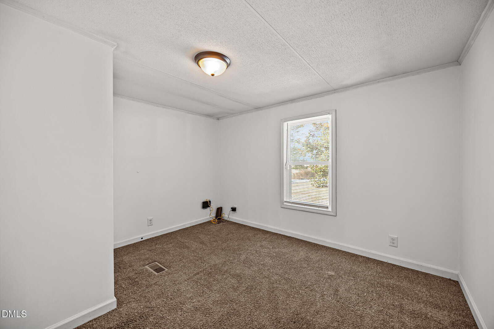 Empty room with white walls, brown carpet, ceiling light, single window letting in daylight, and electrical outlets visible on one wall.