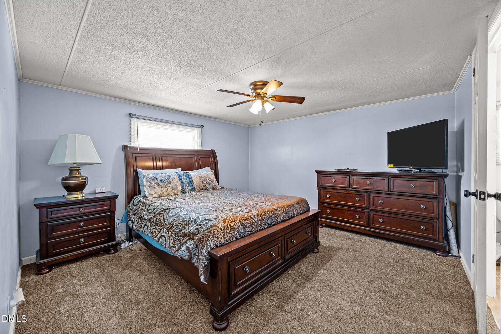 A cozy bedroom features a wooden bed with patterned bedding, matching nightstand and dressers, a lamp, a TV, and a ceiling fan. The walls are light blue and the floor is carpeted. Natural light enters through a window.