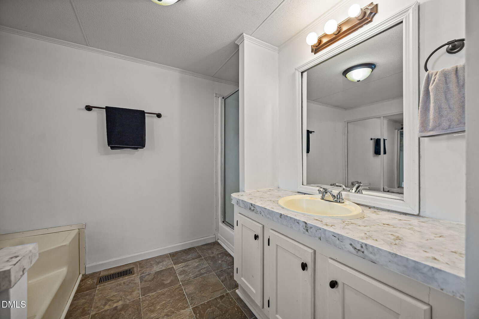 A bathroom with a marble countertop, sink, mirror, vanity lights, towel rack with a dark towel, a bathtub, and tiled floor. The walls are white and there is a ceiling light above.