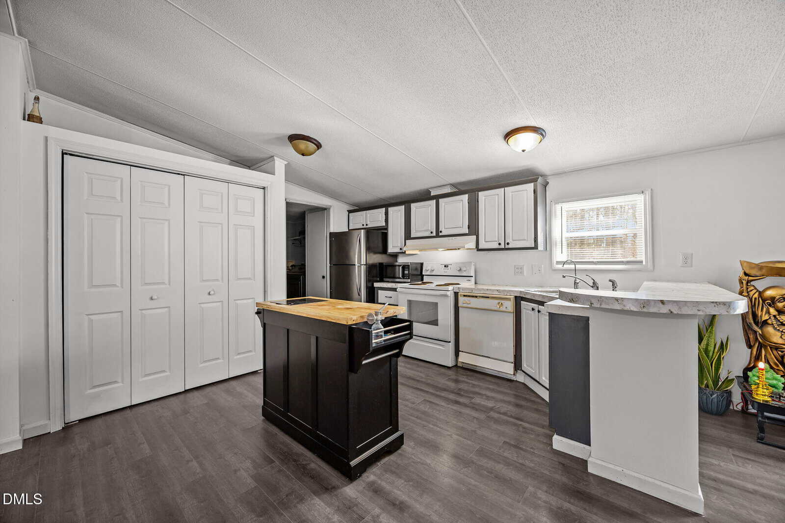 Modern kitchen with white cabinets, stainless steel appliances, a small dark island with a butcher block top, and dark flooring. There are plants and natural light coming from a window above the sink.