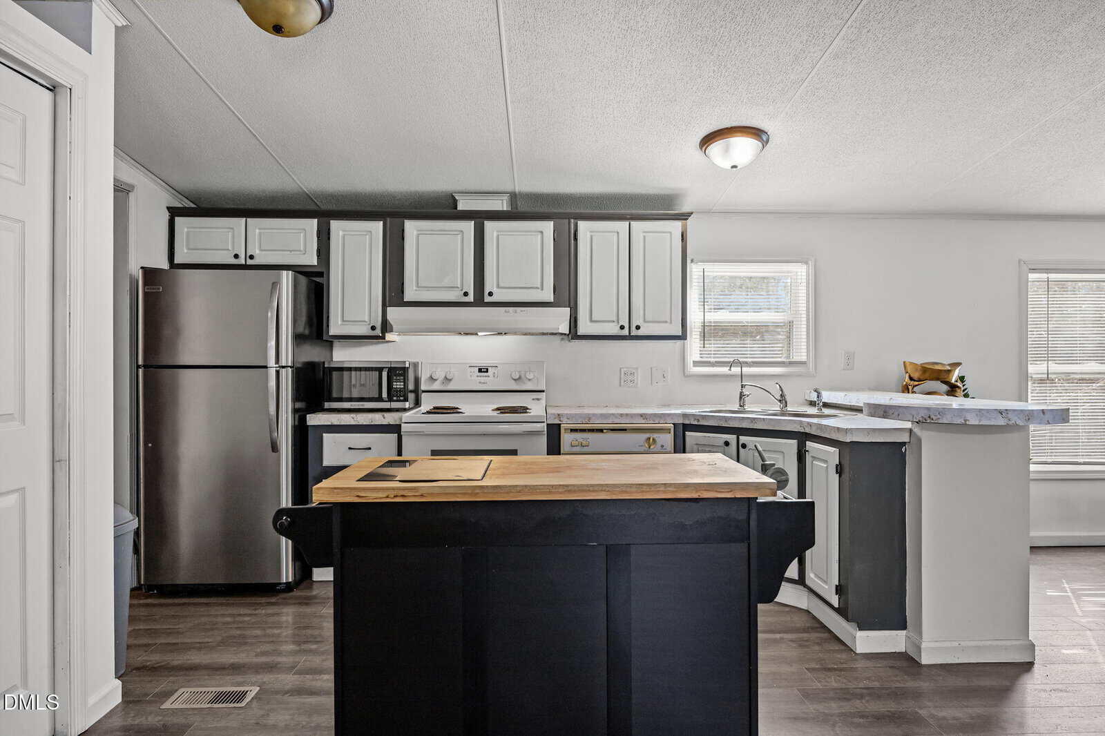 A modern kitchen with dark lower cabinets, white upper cabinets, stainless steel appliances, a central island with a butcher block top, and two windows letting in natural light.