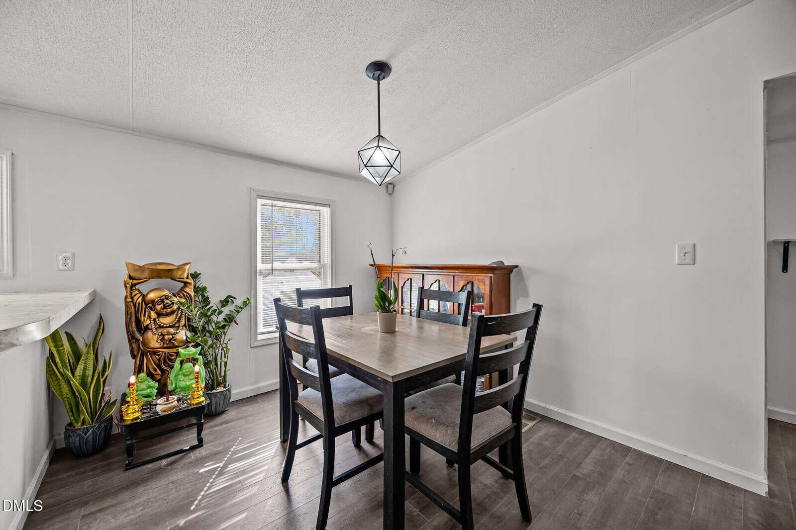 A dining area with a light wood table, four black chairs, a geometric pendant light, plants by a window, a large golden lucky cat statue, and a wooden cabinet against a white wall with dark flooring.